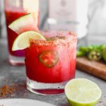 A spicy watermelon margarita with lime slices and jalapeño in a glass rimmed with chili powder, next to a halved lime.